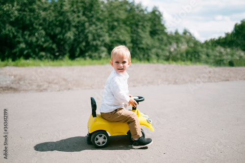 A boy in a white shirt sits on a yellow toy car and looking on the camera. Riding and playing outdoors, outdoor activities.