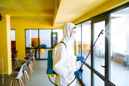 Contractor in a protective suit and mask sprays disinfectants in office before work. Protection against COVID-19 disease. Prevention of spreding pneumonia virus with surfaces. photo