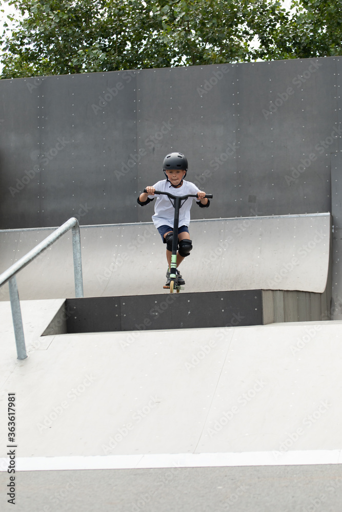 A cute little boy rides a scooter in a skatepark.