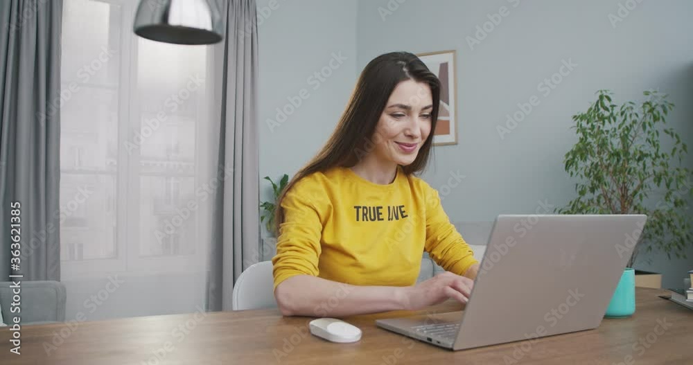 Cheerful beautiful Caucasian dark-haired girl working on laptop indoors at home. Pretty joyful young woman typing and browsing on computer while sitting at table in cozy room. Freelance concept Cheerful beautiful Caucasian dark-haired girl working on laptop indoors at home. Pretty joyful young woman typing and browsing on computer while sitting at table in cozy room. Freelance concept