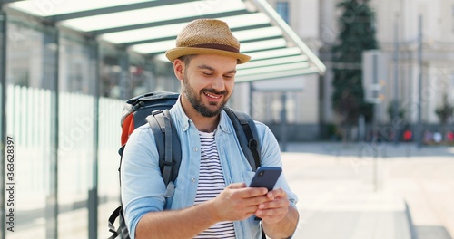 Tablou pe pânză Young handsome Caucasian man in hat with backpack texting message on smartphone and smiling cheerfully at train station