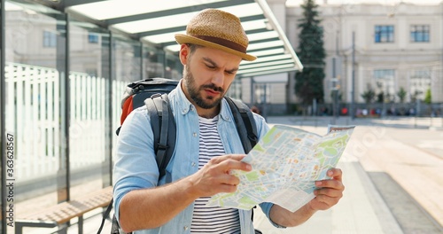 Young handsome Caucasian man tourist in hat with backpack holding city map and planning route. Looking for way in downtown train station. Attractive male traveller holding plan of town at bus stop. photo