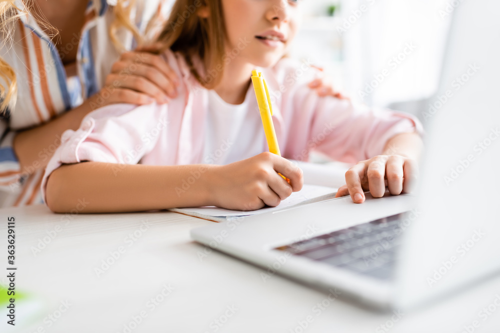 Cropped view of woman embracing kid writing on notebook near laptop on table