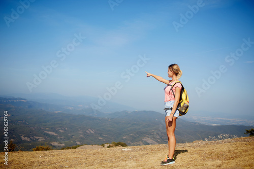 Young woman wanderer points finger on copy space while standing on a mountain hill against amazing view, female traveler enjoying beautiful landscape while taking break during hiking in summer day © BullRun