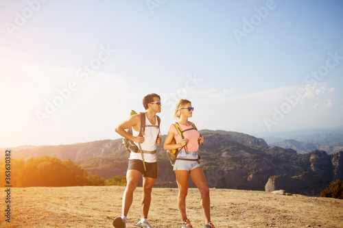 Full length portrait of a young man and woman travelers with rucksacks on backs are admiring wonderful landscape while standing on high mountain in sunny summer day during their joint vacations abroad