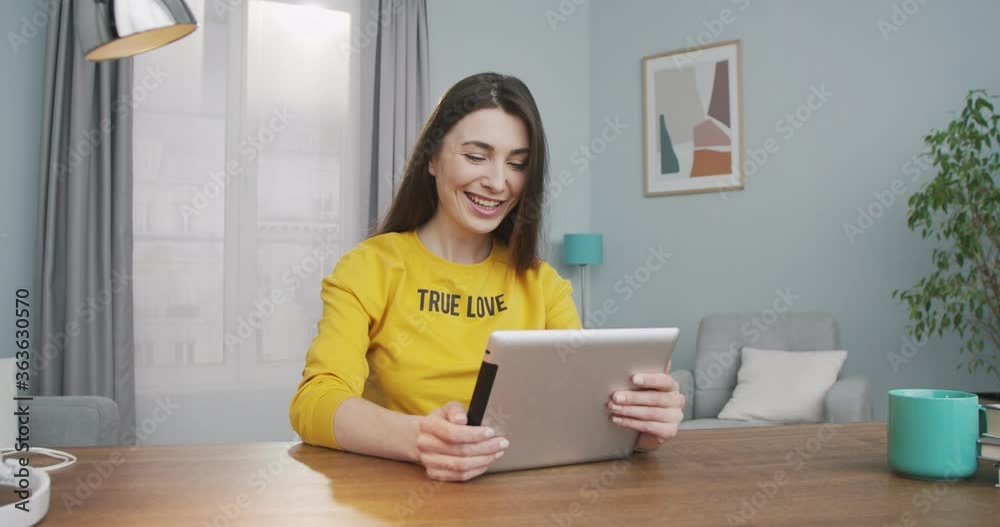 Cheerful pretty girl smiling while video chatting on device indoors. Joyful beautiful Caucasian young woman waving her hand and speaking on video call on tablet in room at home. Conversation concept Cheerful pretty girl smiling while video chatting on device indoors. Joyful beautiful Caucasian young woman waving her hand and speaking on video call on tablet in room at home. Conversation concept