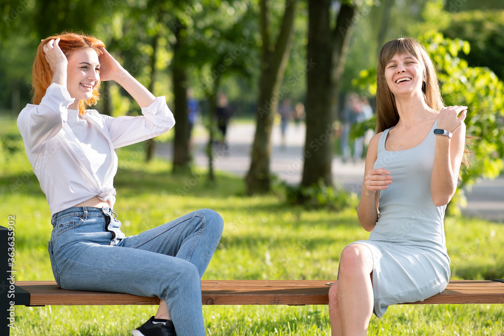 Two young girls friends sitting on a bench in summer park chatting happily having fun.