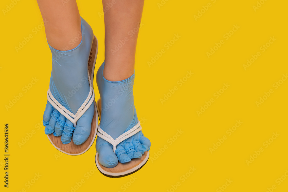 Legs of a girl dressed in rubber blue medical sterile gloves, shod in white flip flops on an orange yellow plain background. A joke of a safe rest on the sea in the sand, protection against a virus Legs of a girl dressed in rubber blue medical sterile gloves, shod in white flip flops on an orange yellow plain background. A joke of a safe rest on the sea in the sand, protection against a virus