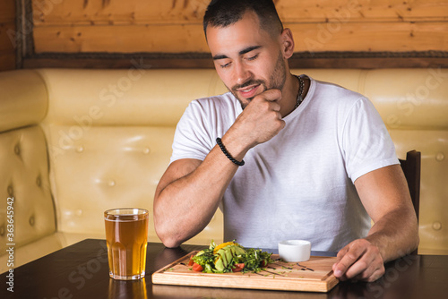 emotional young man sitting at the table, waiting to eat, in front of him a vegetable salad with greens and avocado and meat