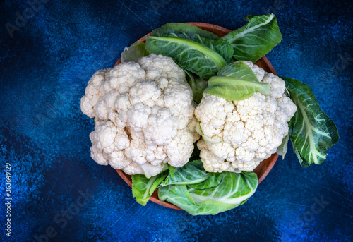 Fresh whole washed cauliflower with leaves in a wooden bowl on a contrasting textured blue background. Concept of natural seasonal fresh vegetables and harvest. Food photo, top view, copy space