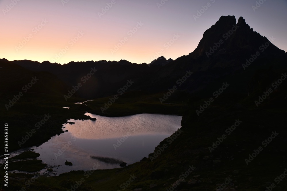 Le soleil dans les Pyrénées françaises. Le ciel devient violet et rose et la montagne se dresse au-dessus d'un lac qui brille. Le paysage est clame et reposant.
