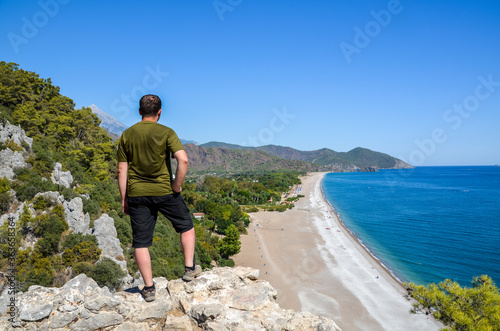 Back view man standing on the rock on the seacoast looking on the breathtaking colorful panoramic view of the Olympos beach in the Cirali in Turkey