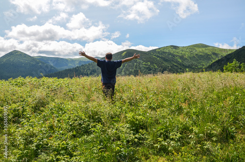 A young man tourist standing at green meadow raised his hands up against backdrop backdrop of Carpathian mountains beautiful scenery landscape