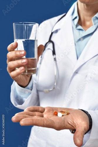 Time to take pills. Cropped shot of a male doctor in medical uniform holding pills and glass of water in front on camera, isolated on blue navy background