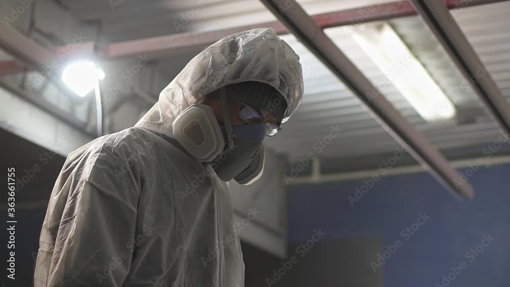 Powder coating of metal products. A man in a White protective suit and a respirator with a spray gun applies a gray powder to an object in the paint chamber. Powder coating of metal products. A man in a White protective suit and a respirator with a spray gun applies a gray powder to an object in the paint chamber.