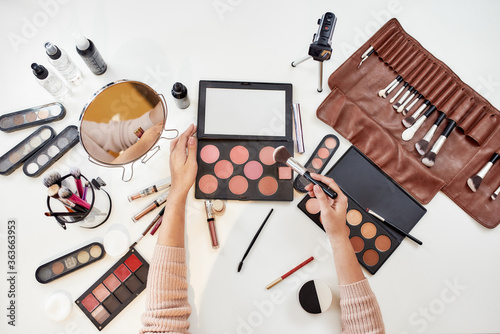 Makeup Inspiration. Top view of young woman, artist doing makeup, sitting at the table. Closeup female hands applying make up. Cosmetic advertising concept