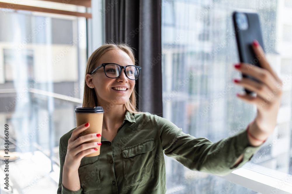 Portrait of woman in formal wear standing holding takeaway coffee in hand and taking selfie on mobile phone in office Portrait of woman in formal wear standing holding takeaway coffee in hand and taking selfie on mobile phone in office