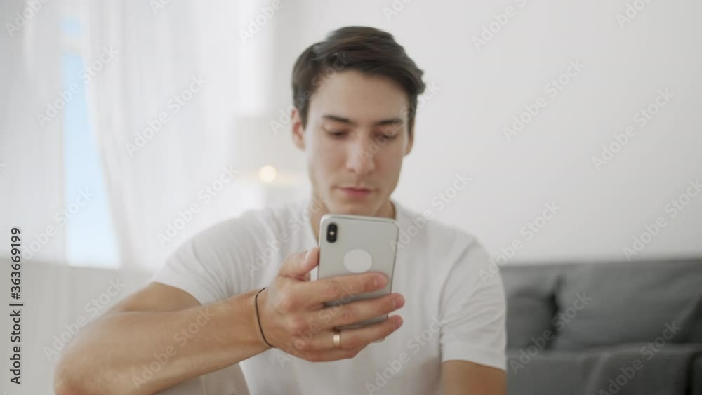 Smiling Young Man Surfing Internet on Phone While Sitting on Exercise Mat During Relaxing After Workout at Home. Smiling Young Man Surfing Internet on Phone While Sitting on Exercise Mat During Relaxing After Workout at Home.
