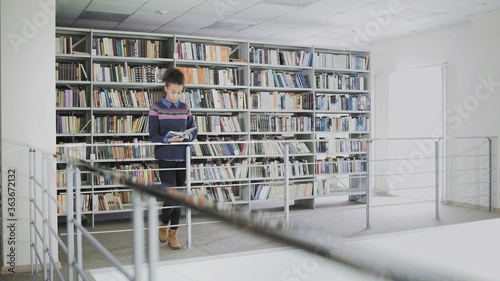 Young pretty african american woman is reading book whlie standing before the shelves in the library