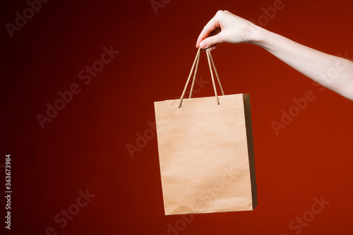 Female hand on a red background holds a paper gift bag. Online delivery and gift concept.
