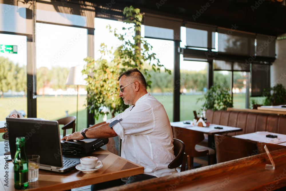 A middle-aged businessman with glasses in a white shirt sits in a modern cafe, works on a laptop and puts business documents in a bag A middle-aged businessman with glasses in a white shirt sits in a modern cafe, works on a laptop and puts business documents in a bag