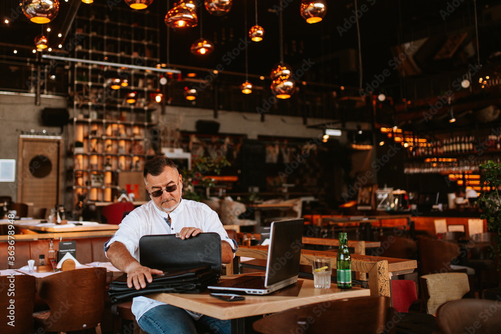 A middle-aged businessman with glasses in a white shirt sits in a modern cafe, works on a laptop and puts business documents in a bag