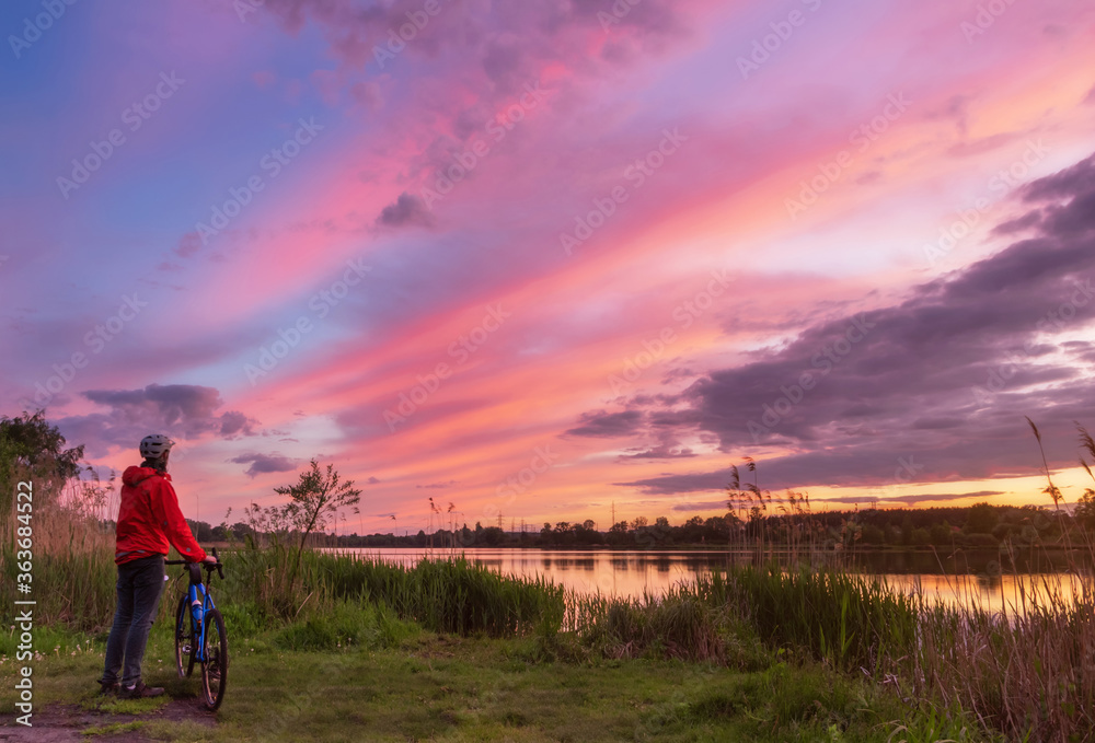 Cyclist on the coast of the river at sunset in summer. Landscape with man on gravel bike and colorful sky with clouds. Sport and travel concept.