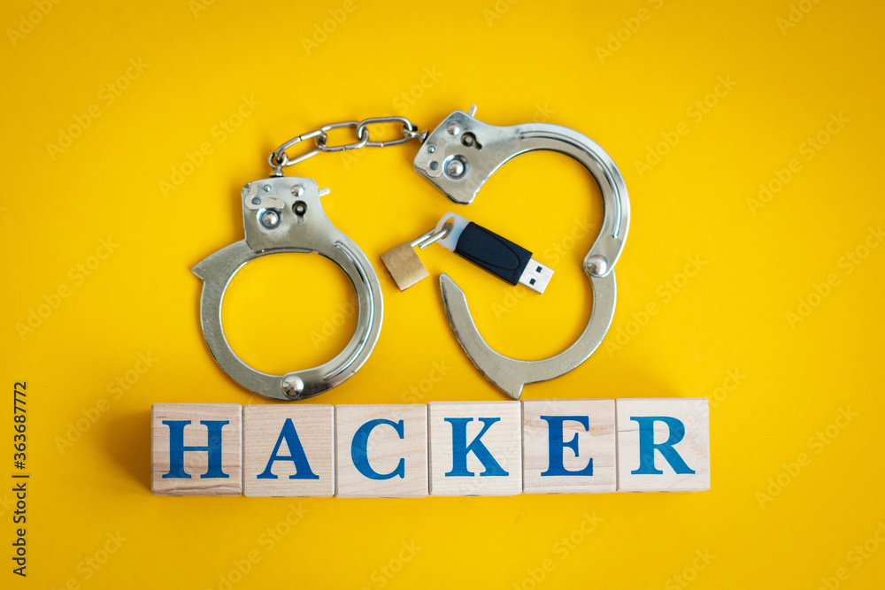 Handcuffs, a flash drive with an open lock and the word HACKER from cubes on a yellow background. The concept of the punishment for cybercrime and hacking information. Handcuffs, a flash drive with an open lock and the word HACKER from cubes on a yellow background. The concept of the punishment for cybercrime and hacking information.