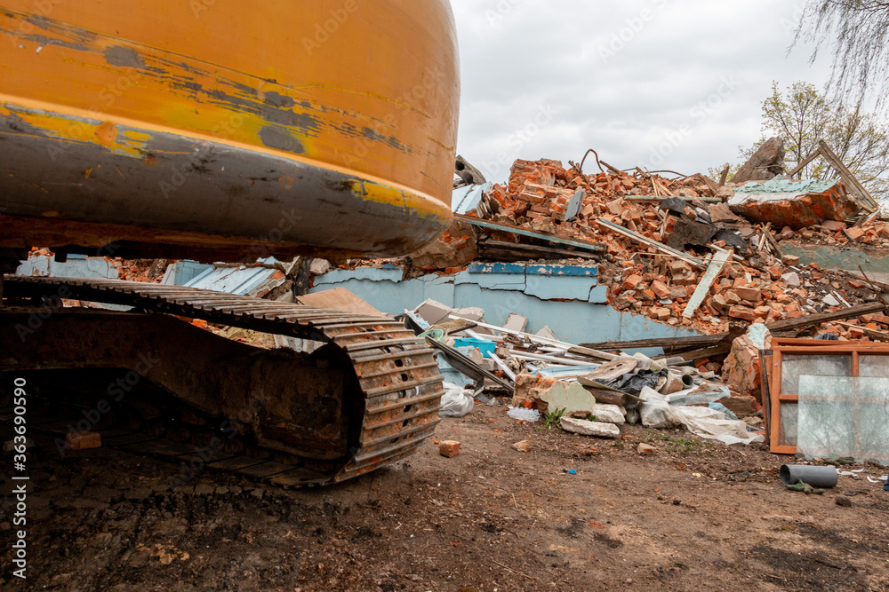 Fototapeta premium Excavator caterpillar on the background of a destroyed, brick, residential building. Demolition of an old house with an excavator.