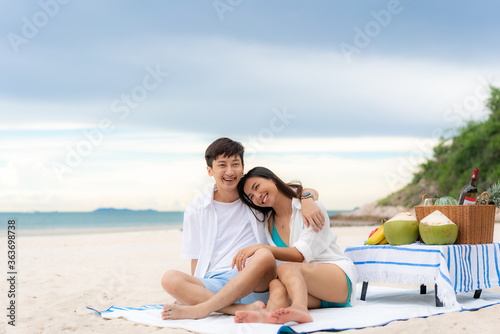 Asian young couple sitting on the picnic blanket and relax in the white sand beach and near sea with tropical fruit in background. Summer, holidays, vacation and happy people in Thailand concept © ake1150