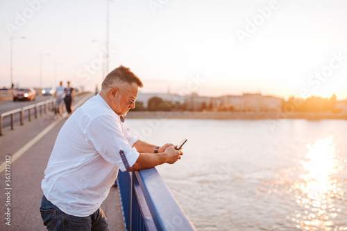 A middle-aged caucasian businessman with glasses, in a white shirt, is typing a message on the phone leaning against the bridge railing.