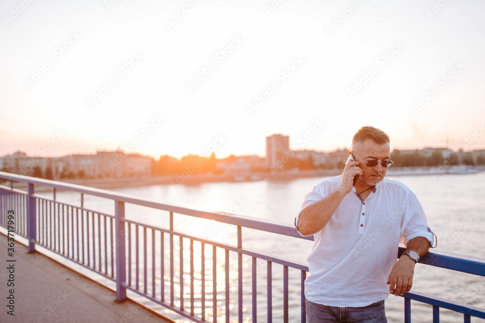 A middle-aged caucasian businessman with glasses, in a white shirt, talking on the phone leaning against the bridge railing