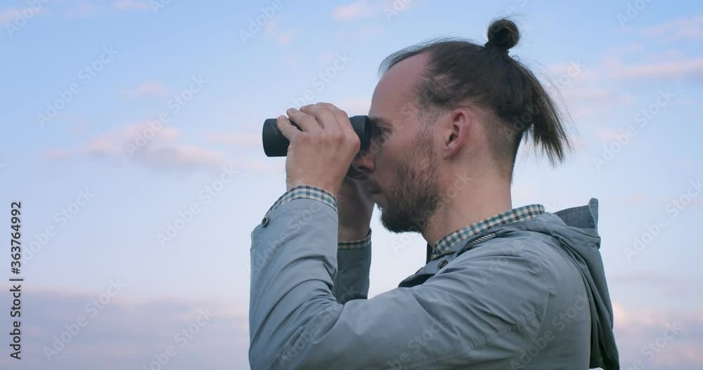 Side portrait of hipster bearded man looking into binoculars afar handheld camera slow motion. Authentic man with eastern hairstyle holding glasses outdoors copy text space. Travel adventure equipment Side portrait of hipster bearded man looking into binoculars afar handheld camera slow motion. Authentic man with eastern hairstyle holding glasses outdoors copy text space. Travel adventure equipment