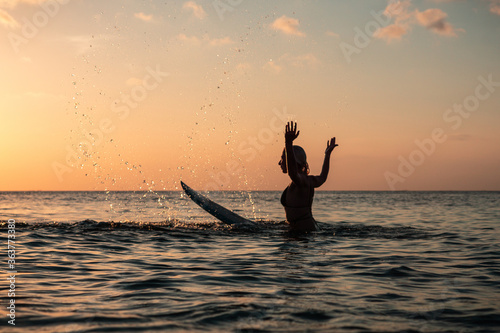 Portrait from the water of surfer girl with beautiful body on surfboard splashes water in the ocean at colourful sunset time