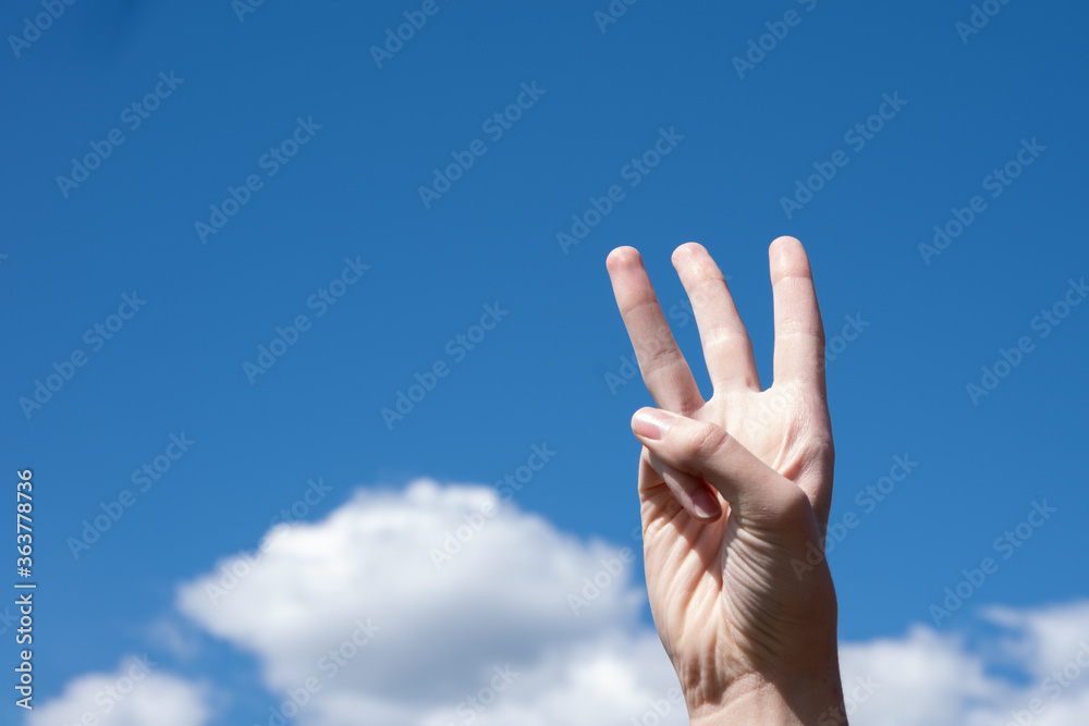 Close-up gesture of a woman's hand showing three fingers isolated on a background of blue sky with clouds Close-up gesture of a woman's hand showing three fingers isolated on a background of blue sky with clouds