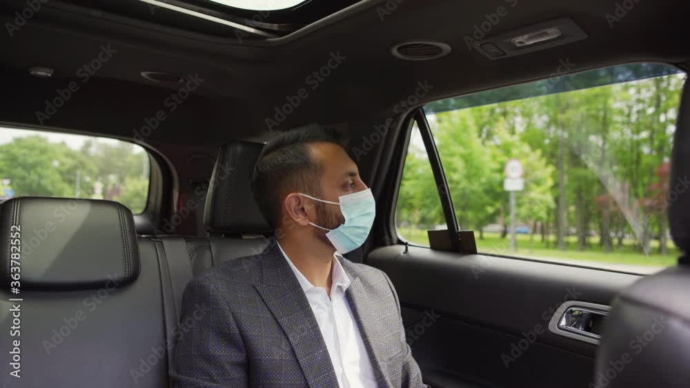 Tilt up of young Middle Eastern businessman in suit and medical face mask typing on laptop and looking thoughtfully through window sitting in backseat of car Tilt up of young Middle Eastern businessman in suit and medical face mask typing on laptop and looking thoughtfully through window sitting in backseat of car