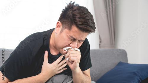 Asian man with seasonal infections Cold Blowing His Nose and sneezing into Tissue with headache lying on sofa with high fever and a flu