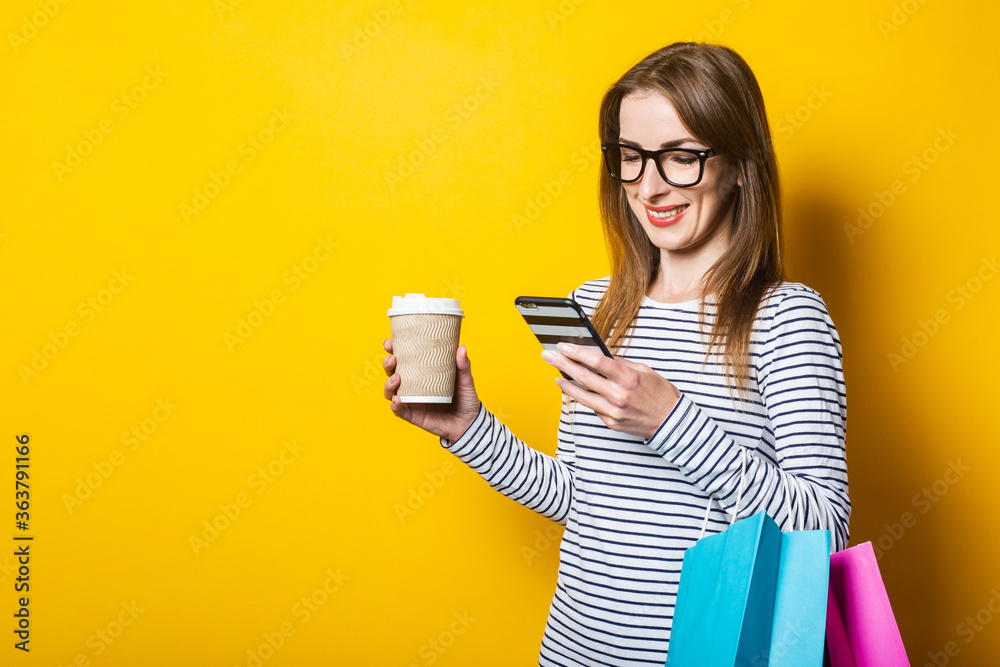 Smiling young girl looking at the phone, with a paper cup with coffee and with shopping bags on a yellow background.
