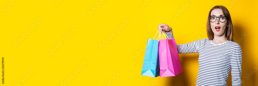 Surprised shocked young woman holds shopping bags on a yellow background. Banner