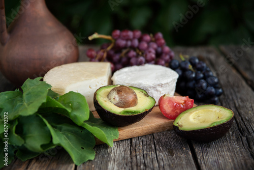 Clay jug, grapes, heads of cheese, avocado, tomato and arugula on a wooden background.