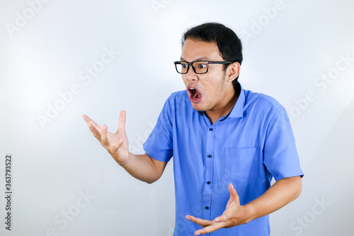 Young Asian Man wear blue shirt is funny angry face with shouting and pointing finger at camera isolated over white background © Reezky