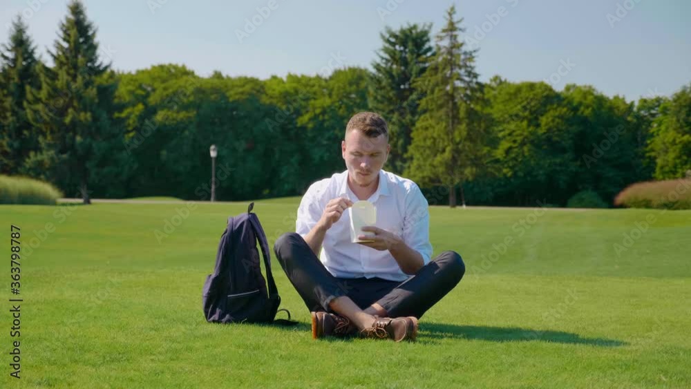 Man eats vegetarian lunch with an ecological package, shows like and smiles. Guy sits in the park on the green grass eats salad with tomatoes, cucumbers and lettuce. Wooden fork. Paper packaging Man eats vegetarian lunch with an ecological package, shows like and smiles. Guy sits in the park on the green grass eats salad with tomatoes, cucumbers and lettuce. Wooden fork. Paper packaging