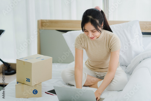 Young Asian woman in casual outfit working from home sitting on bed using computer laptop and checking package box before send to customer in modern bedroom