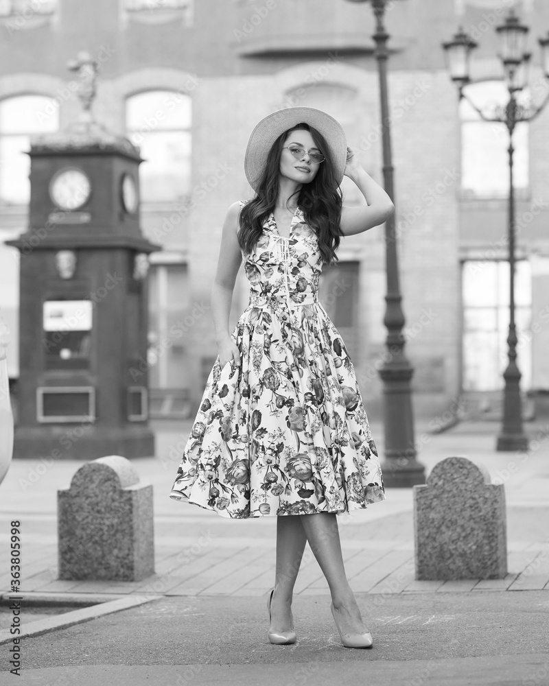 Fototapeta premium Young woman in colorful dress walking city street on a sunny summer day. Full length portrait. Elegant lady with long wavy hair. Girl in sunglasses