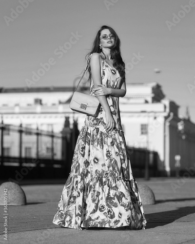 Elegant woman wearing white and blue colorful summer dress and sunglasses and walkig city street. Evening full length portrait. Fashionable female model posing outdoors photo