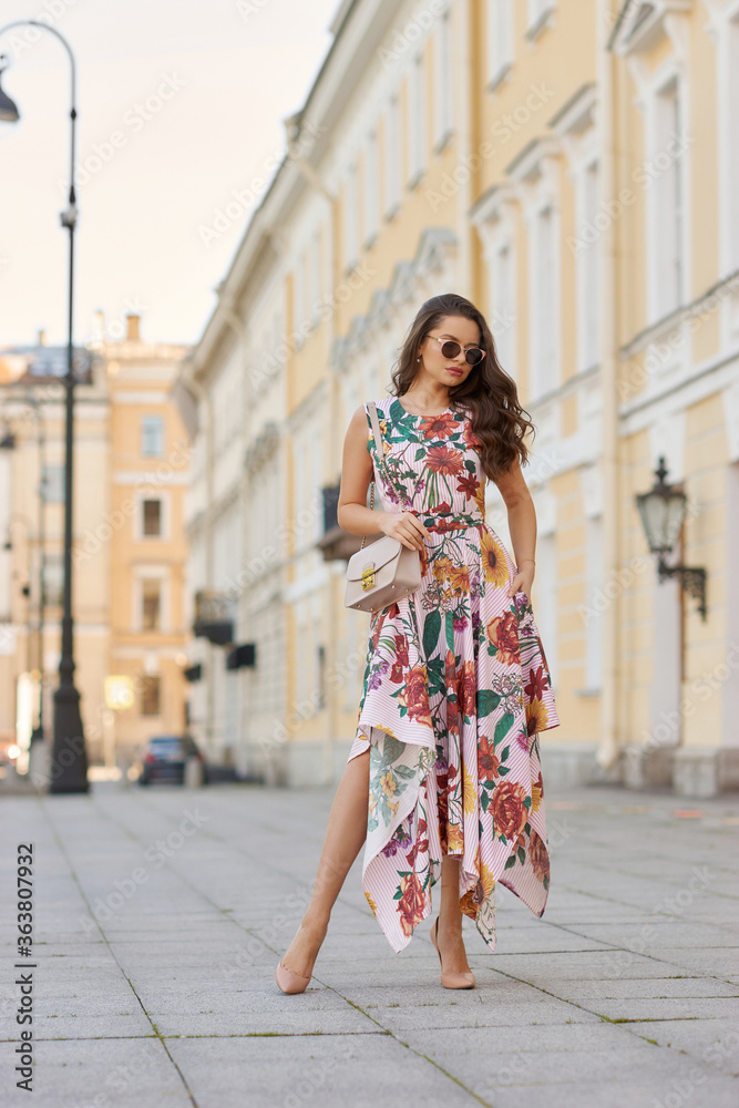 Fototapeta premium Young beautiful elegant tall slim woman with natural makeup and wavy brunete hair wearing colorful dress and sunglassses walking in the city on a summer day and holding handbag