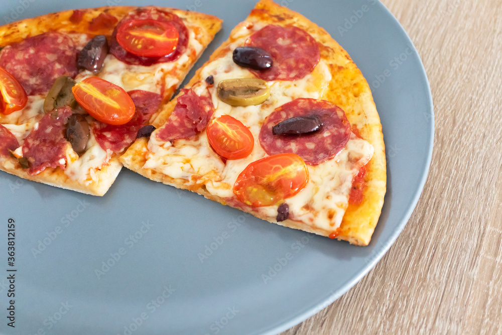 Two pieces of homemade cheese pizza with salami, olives and cherry tomato on gray plate on wooden table, selective focus. Hot tasty pepperoni Two pieces of homemade cheese pizza with salami, olives and cherry tomato on gray plate on wooden table, selective focus. Hot tasty pepperoni