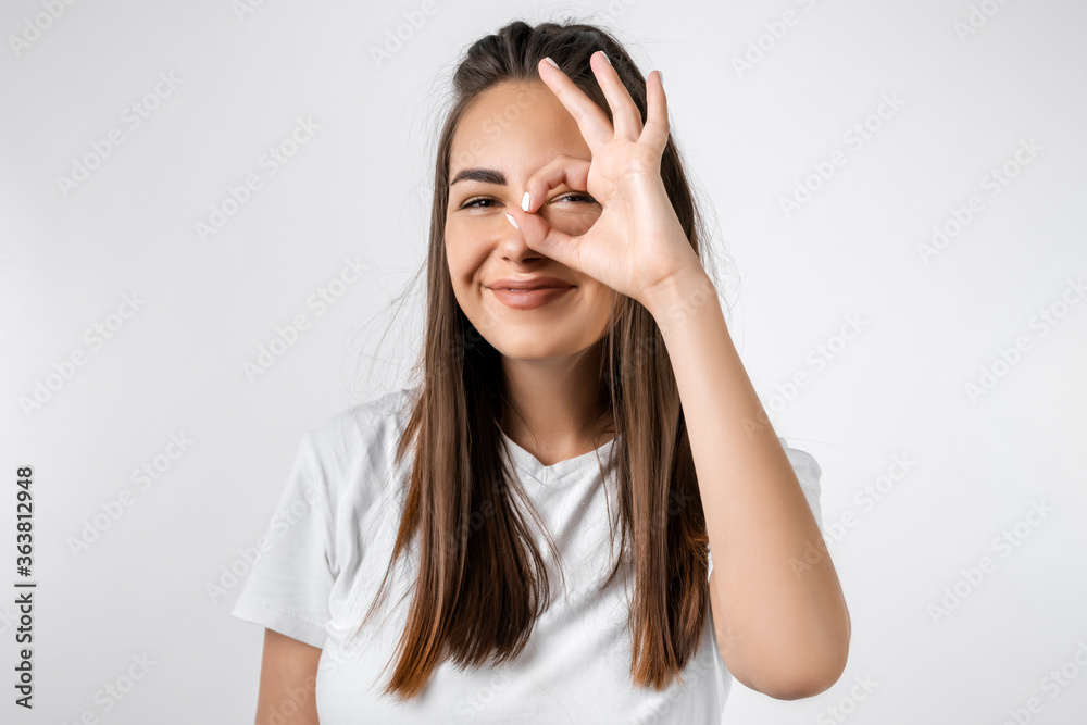 Close up portrait of carefree modern girl with long chestnut hairs, looking at the camera through fingers in OKAY gesture. Face expressions, emotions, and body language Close up portrait of carefree modern girl with long chestnut hairs, looking at the camera through fingers in OKAY gesture. Face expressions, emotions, and body language