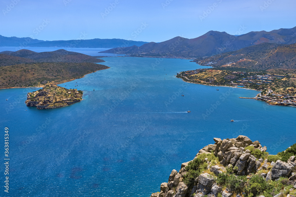 View of harbor between Spinalonga island and peninsula, known as Kalydon, and Plaka village from above and rocky slope in foreground, Crete, Greece.