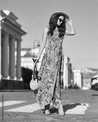 Young beautiful elegant tall slim woman with natural makeup and wavy brunete hair wearing colorful dress, salty hat and sunglassses walking in the city on a summer day and holding handbag photo